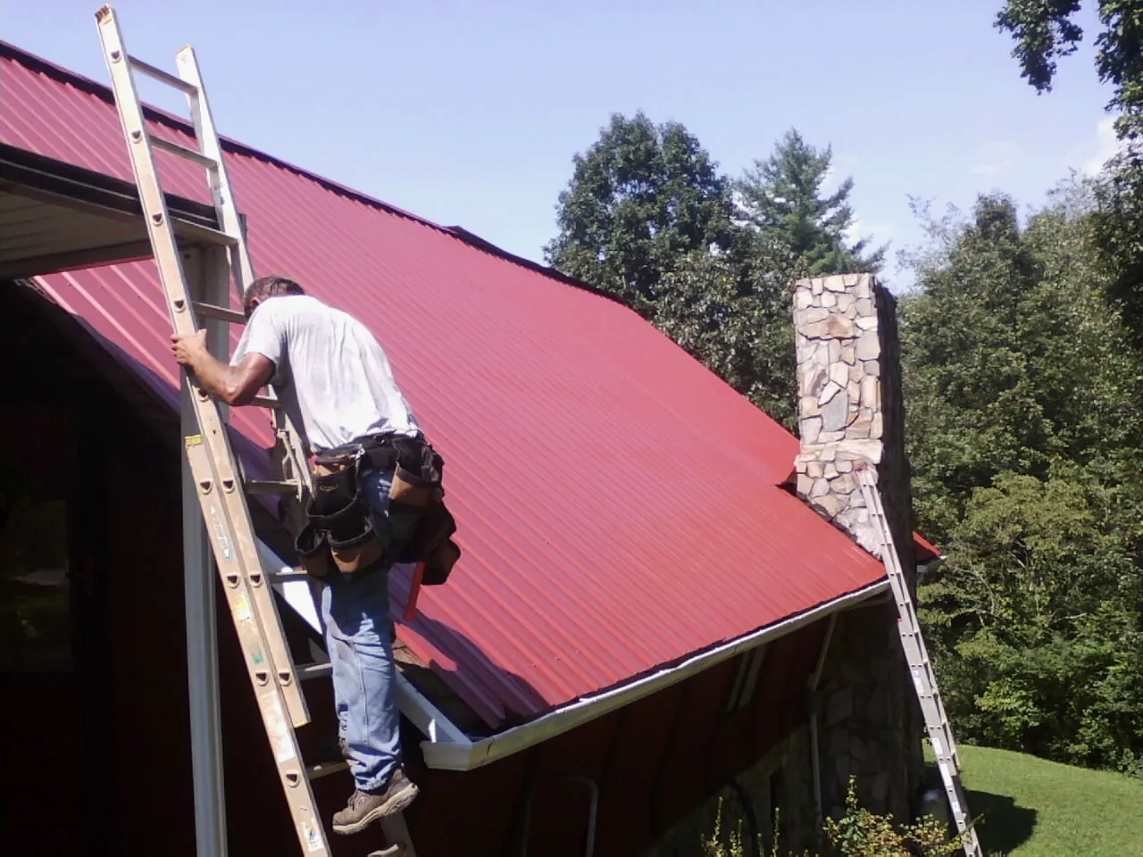 Roofer on a ladder beside a red metal roof and stone chimney — Soffit & Fascia Repair by Roofing Brothers in Scotland